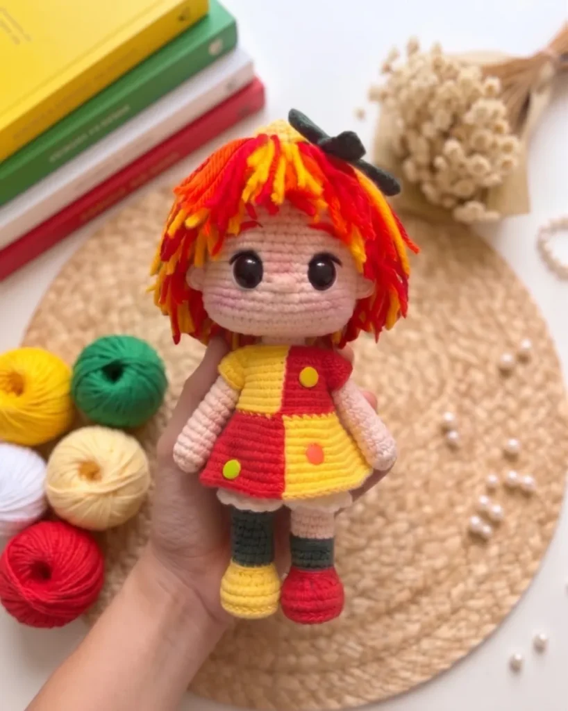 Boneca Emília de amigurumi segurada na palma da mão, com novelos vermelho, amarelo e verde ao fundo